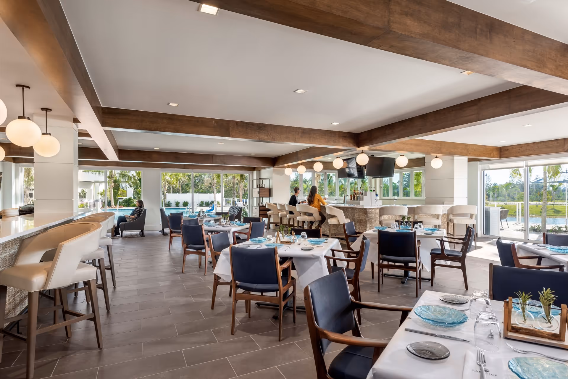 Open modern dining room with set tables, bar seating, and large windows overlooking a pool and outdoor area.