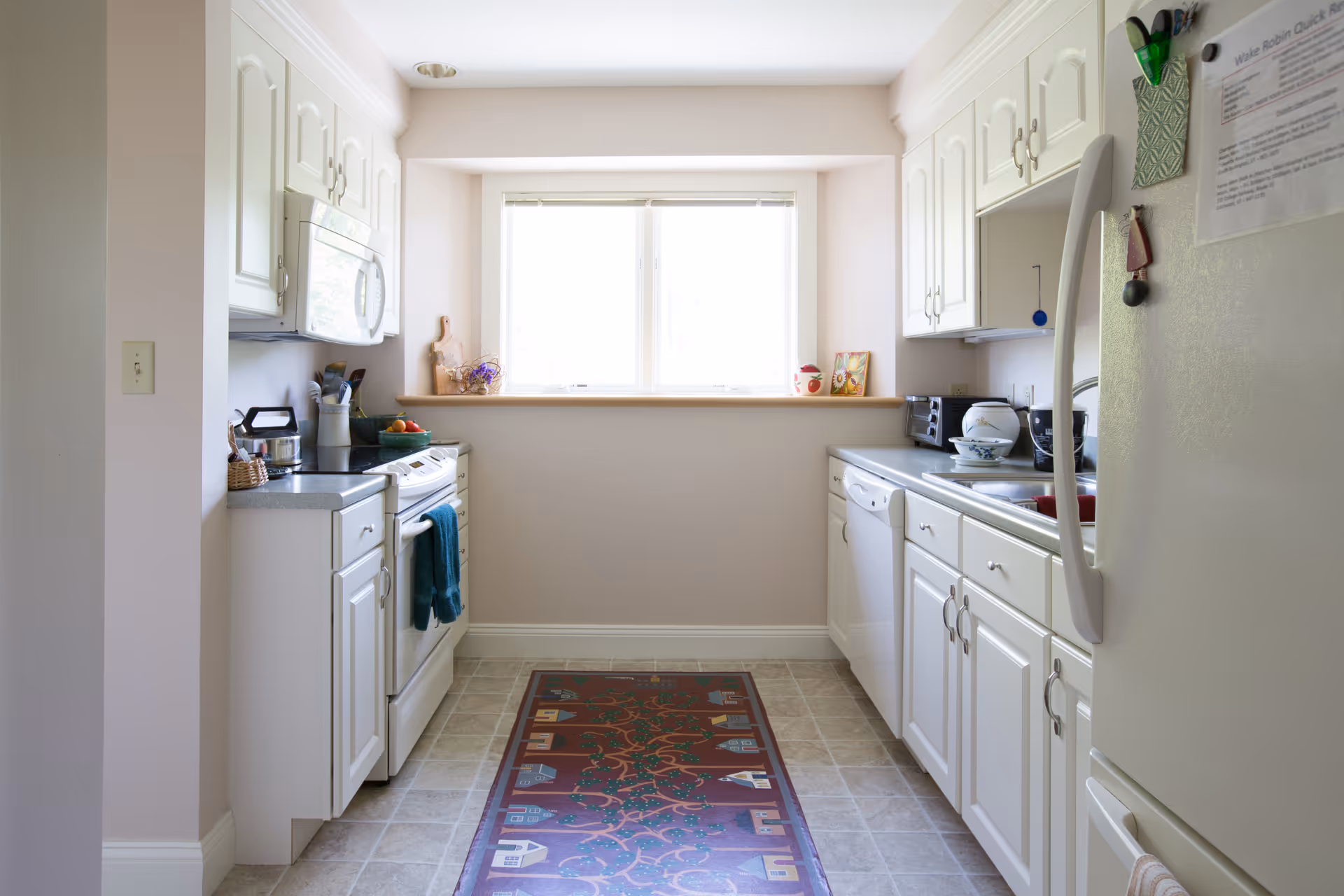 A bright kitchen with white cabinets and appliances on both sides. There is a window at the end letting in natural light. The floor has a decorative rug with a house pattern. Various kitchen items such as a toaster, bowls, and utensils are visible on the countertops.
