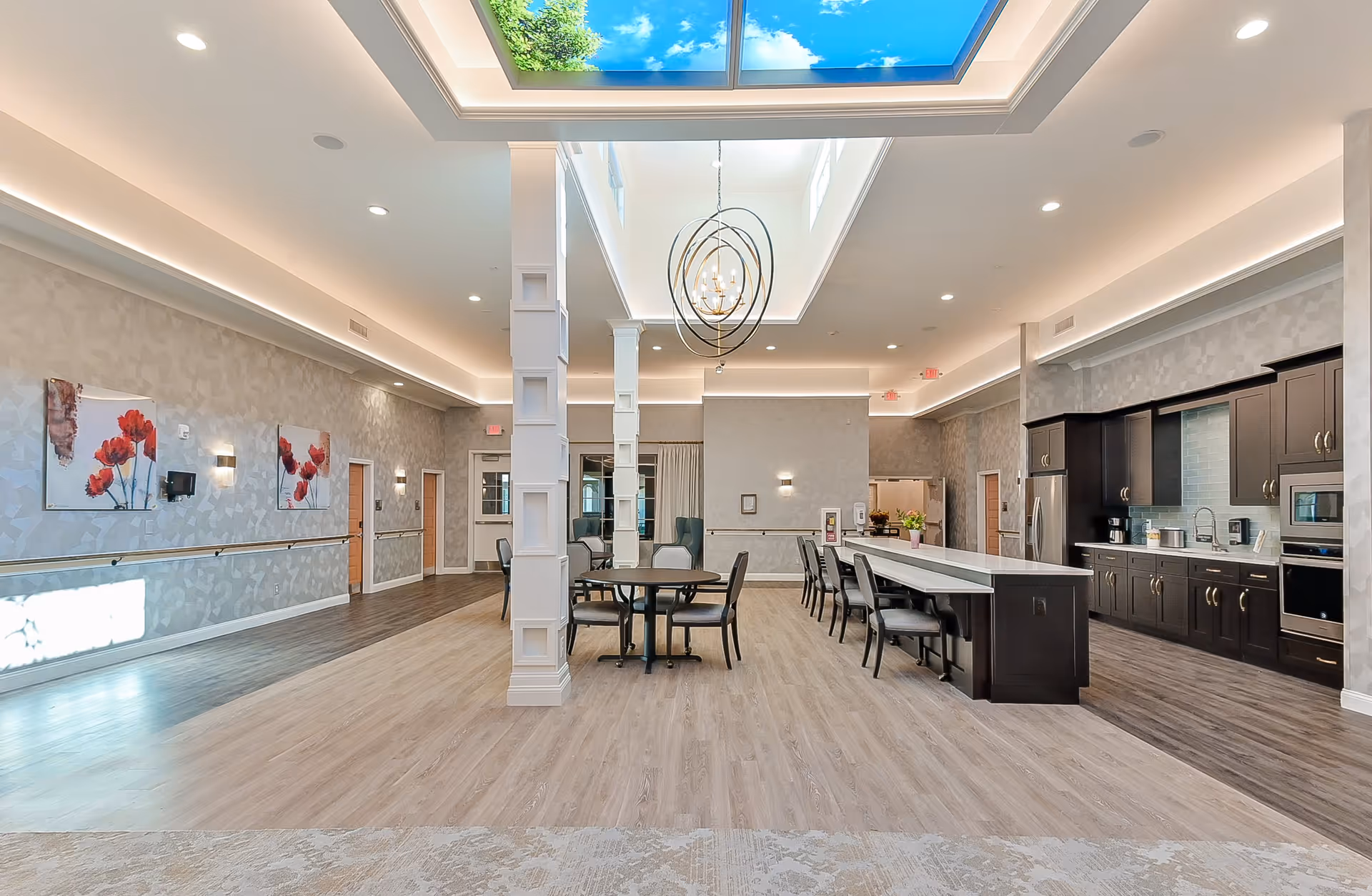 A spacious and modern interior room featuring a kitchen area with dark cabinets, stainless steel appliances, and a long island with chairs. Adjacent to the kitchen is a dining area with tables and chairs. The room has light wood flooring, soft wall lighting, decorative wall art with red flowers, and a large skylight ceiling with a contemporary chandelier.