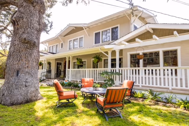 Outdoor seating area with four orange cushioned chairs arranged around a round table on a grassy lawn in front of a two-story beige building with a white railing and large windows.