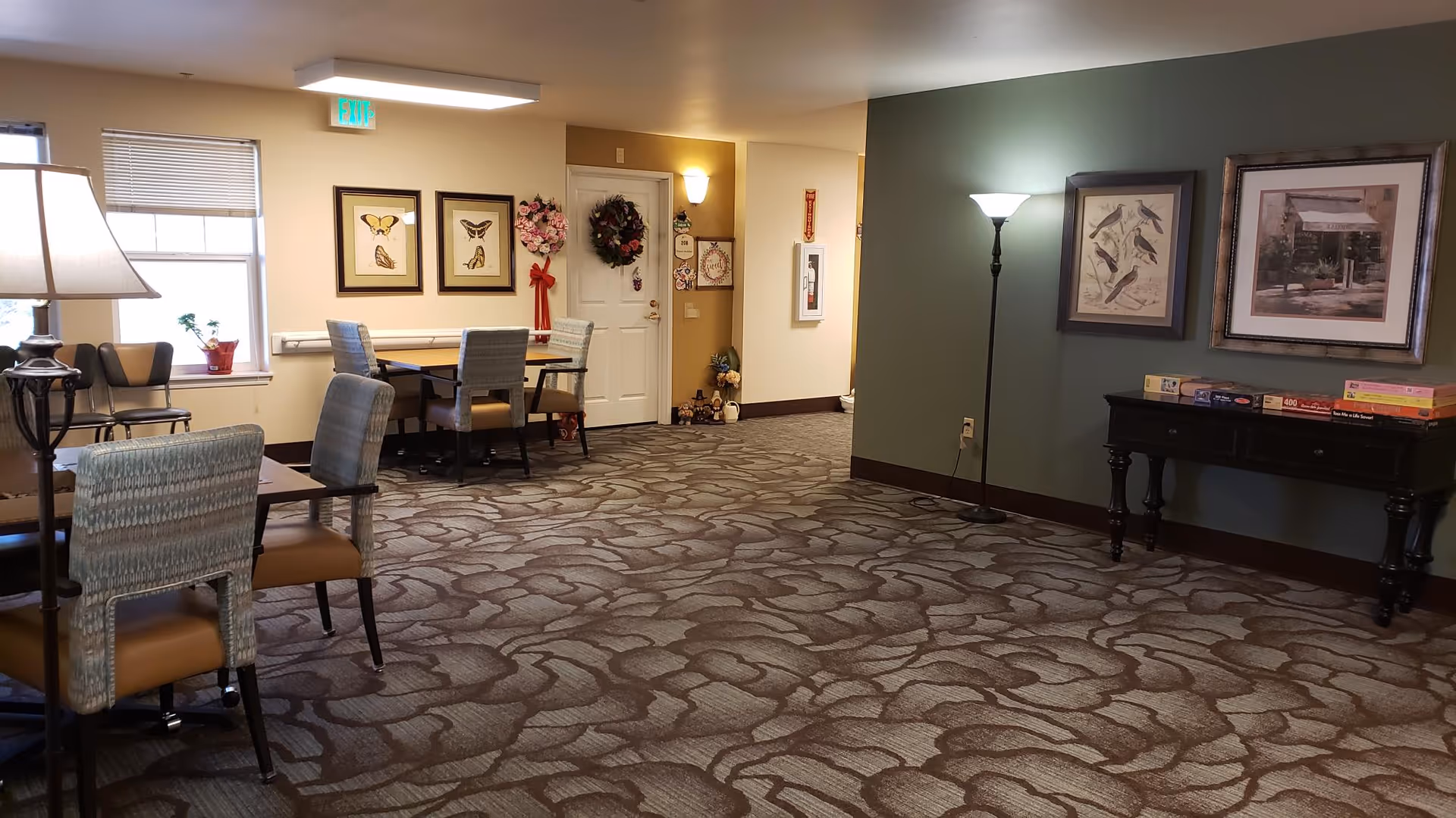 A common area in a senior living facility with patterned carpet, several tables and chairs, framed artwork on the walls, a floor lamp, and a small table with board games on it. There are wreaths and decorations near a white door, and a window with blinds partially open letting in natural light.