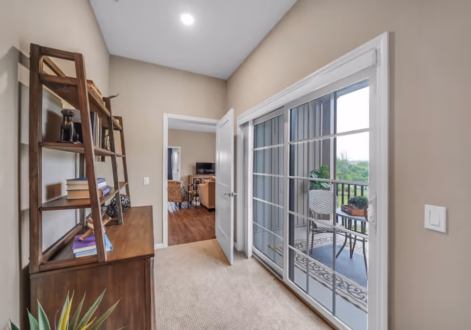 A hallway with beige walls and carpeted floor leading to a living room with wooden flooring. On the left side of the hallway is a wooden shelf with books and decorative items. On the right side, there are large sliding glass doors opening to a balcony with a chair, small table, and potted plants. The living room visible through the doorway has a TV and seating area.