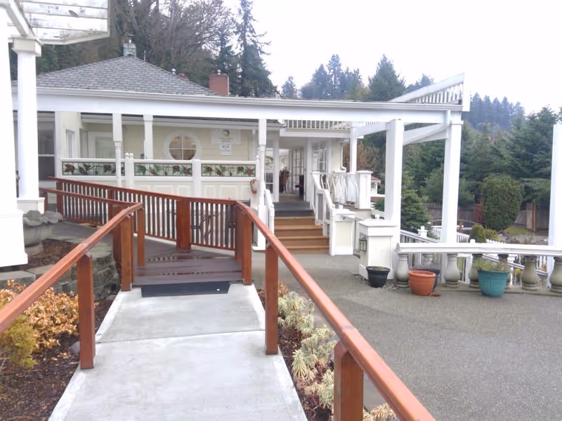Covered front entrance with a wooden wheelchair ramp and steps leading to a white house-style building with railings and potted plants.