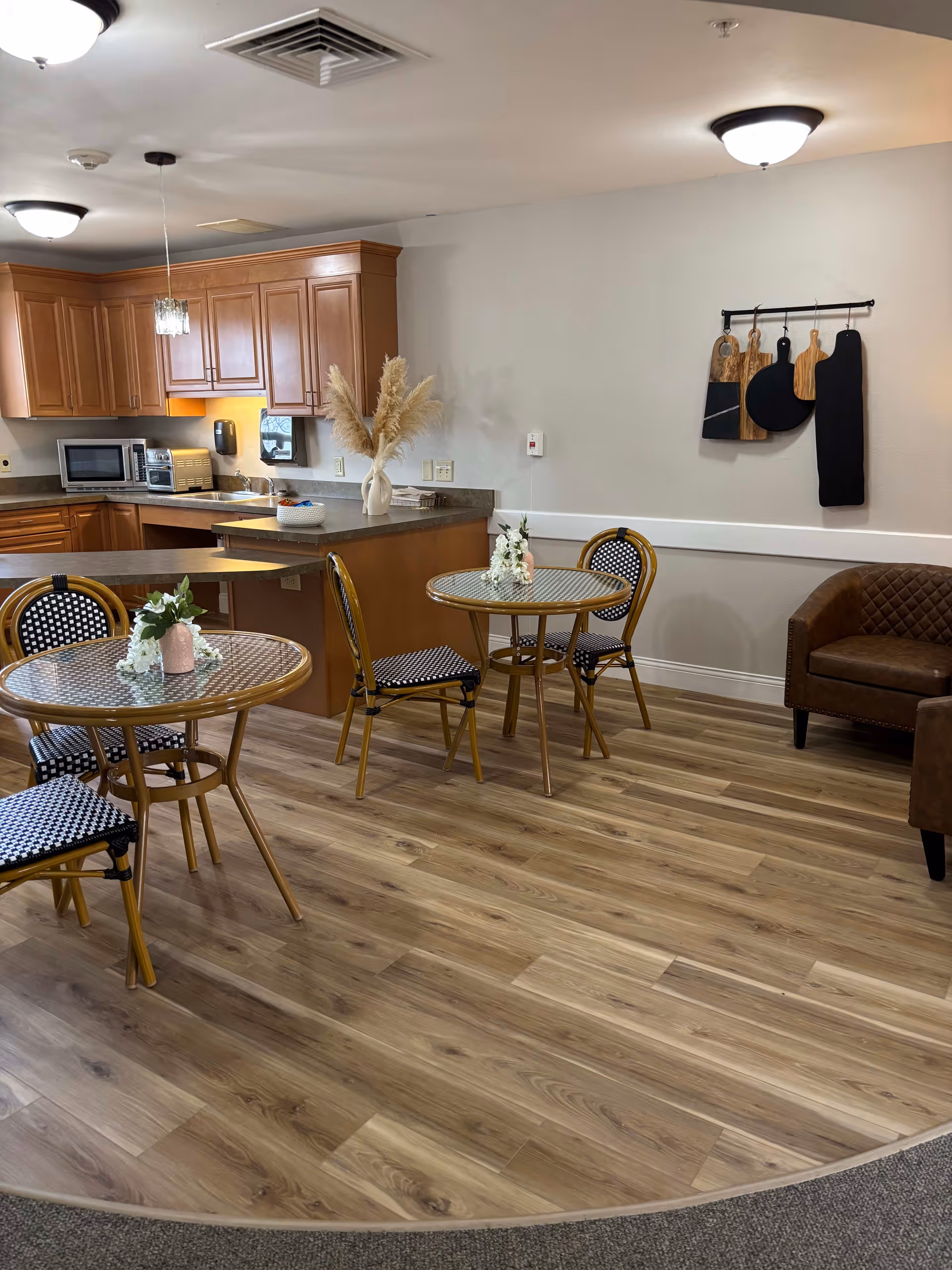 A cozy dining area with two round tables, each surrounded by three chairs with black and white patterned seats. The room features wood flooring and a kitchen area with wooden cabinets, a microwave, and a toaster oven. Decorative items include a vase with pampas grass on the kitchen counter, small flower arrangements on the tables, and a wall-mounted rack holding various cutting boards. There is also a brown upholstered chair in the corner.