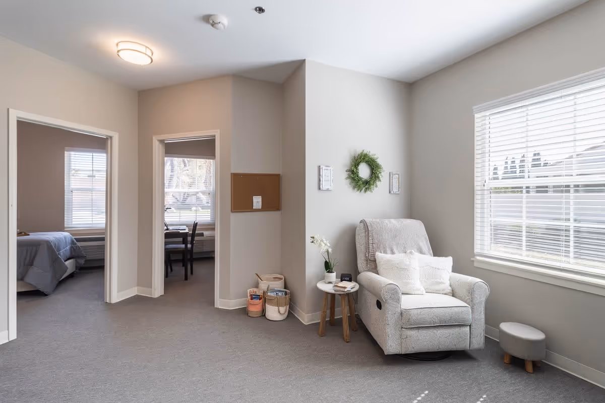 A bright and cozy interior space with a comfortable armchair and small side table near a large window with blinds. The walls are light-colored with minimal decorations including a green wreath and two small framed pictures. Two doorways lead to other rooms, one showing a bed and the other a desk with chairs near a window. The floor is carpeted in a neutral tone.