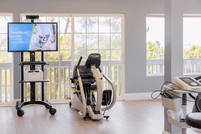 A bright room with large windows showing an exercise machine and a monitor on a stand displaying information about OmniVersa, a versatile, powerful, and accessible device. The room has light-colored walls and wood flooring.