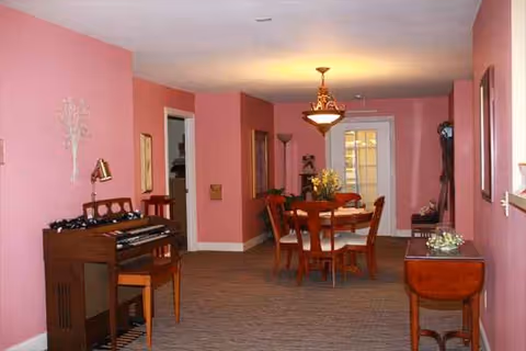 Pink-walled dining room with round table, chairs, piano, and decorative furniture.