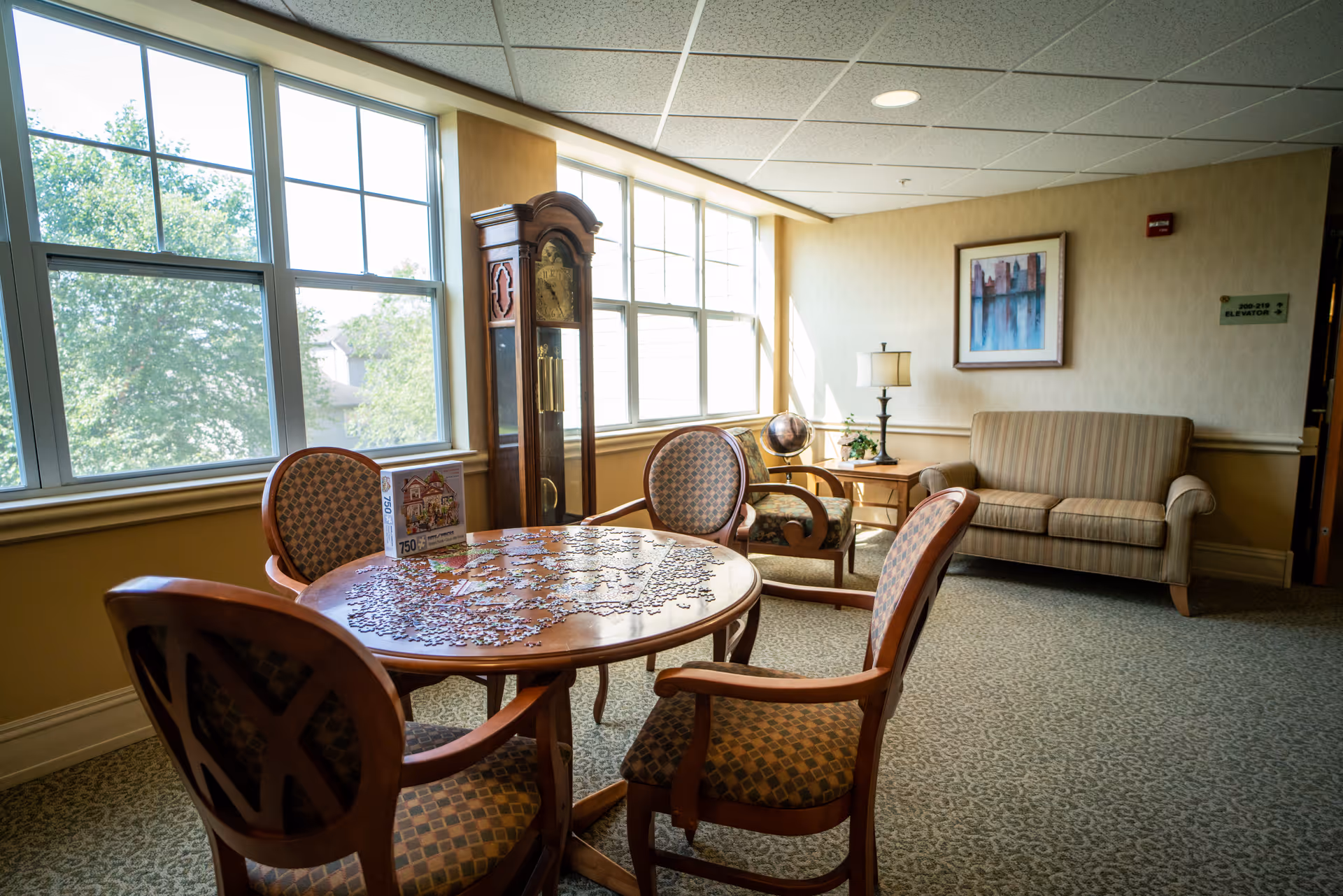 Sunlit common room with a round table covered in a jigsaw puzzle, upholstered chairs, a grandfather clock, and a sofa by large windows.