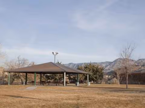 Open-air pavilion with picnic tables beneath a shelter, set in a grassy field with mountains.