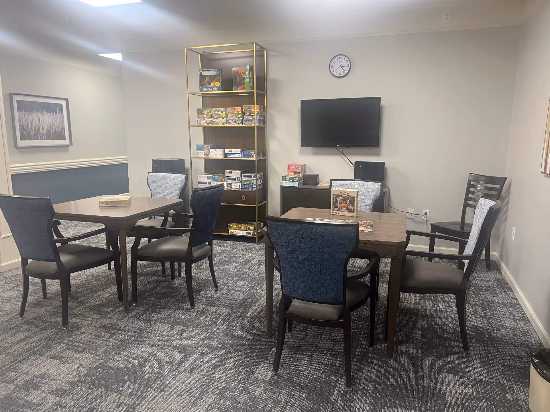 A senior living facility activity room with two square tables surrounded by chairs. On the tables are puzzle boxes and partially completed puzzles. A tall shelving unit against the wall holds various puzzle boxes. A flat-screen TV is mounted on the wall above a cabinet. The room has carpeted flooring, neutral-colored walls, a clock on the wall, and framed artwork.