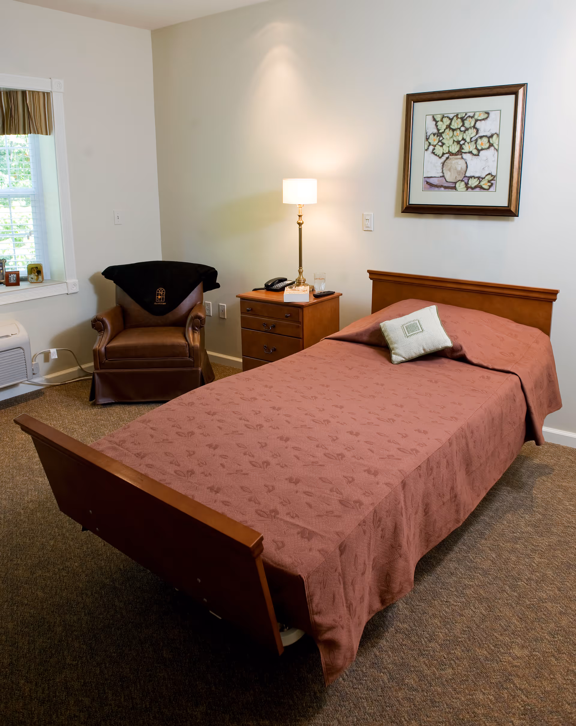 A neatly made single bed with a brown patterned bedspread and a small decorative pillow in a room. Next to the bed is a wooden nightstand with a lamp, a phone, a glass of water, and a box of tissues. There is a brown leather armchair with a black throw draped over the back near a window with white blinds and a valance. A framed floral painting hangs on the wall above the bed.