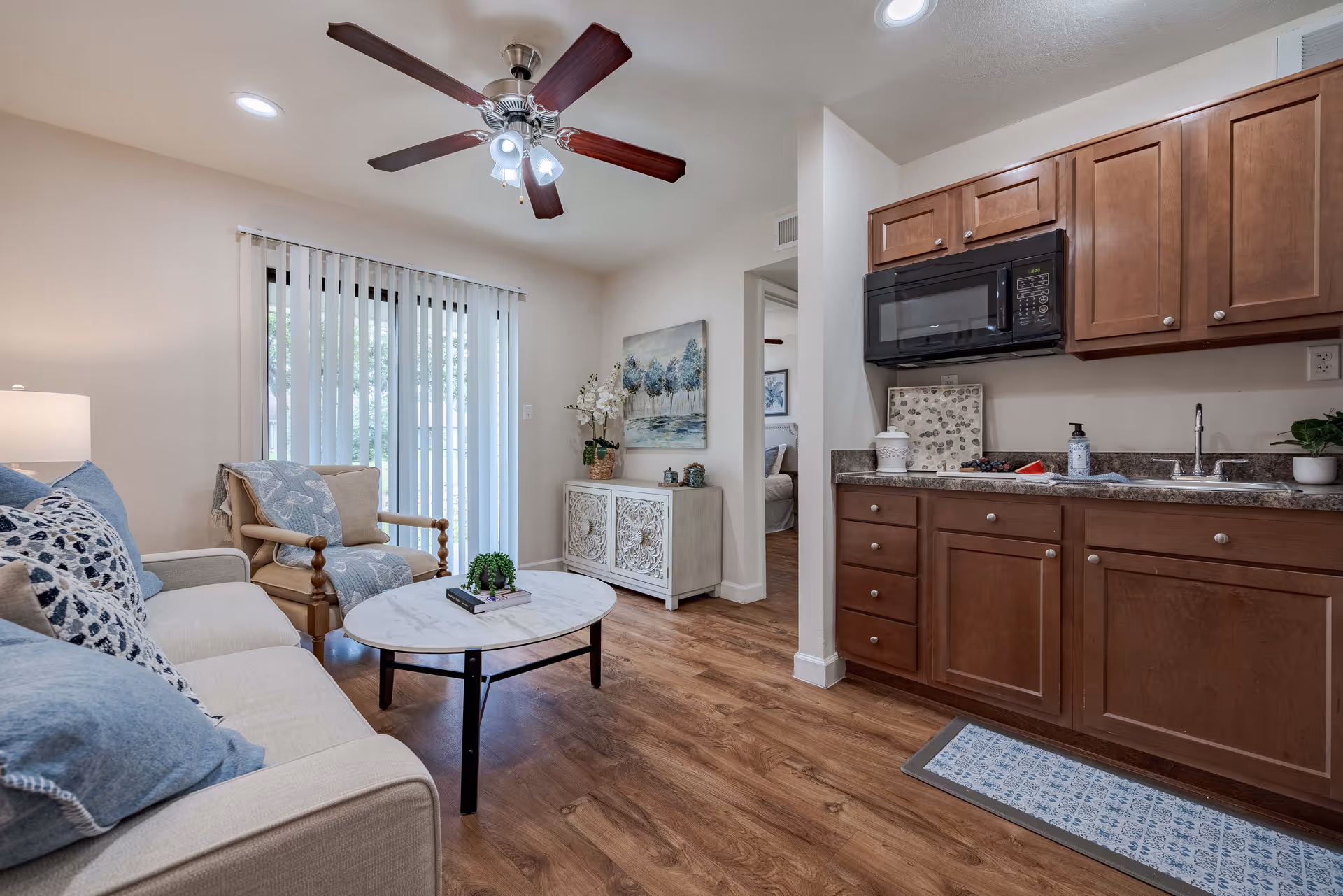 A cozy living room and kitchen area in a retirement community apartment. The living room features a beige sofa with blue and patterned pillows, a wooden armchair with a blue throw, a round marble coffee table with a small plant and books, and a ceiling fan with lights. The kitchen has wooden cabinets, a black microwave, a sink, and a countertop with a soap dispenser and decorative items. There is a sliding glass door with vertical blinds letting in natural light, and a glimpse of a bedroom through an open doorway.