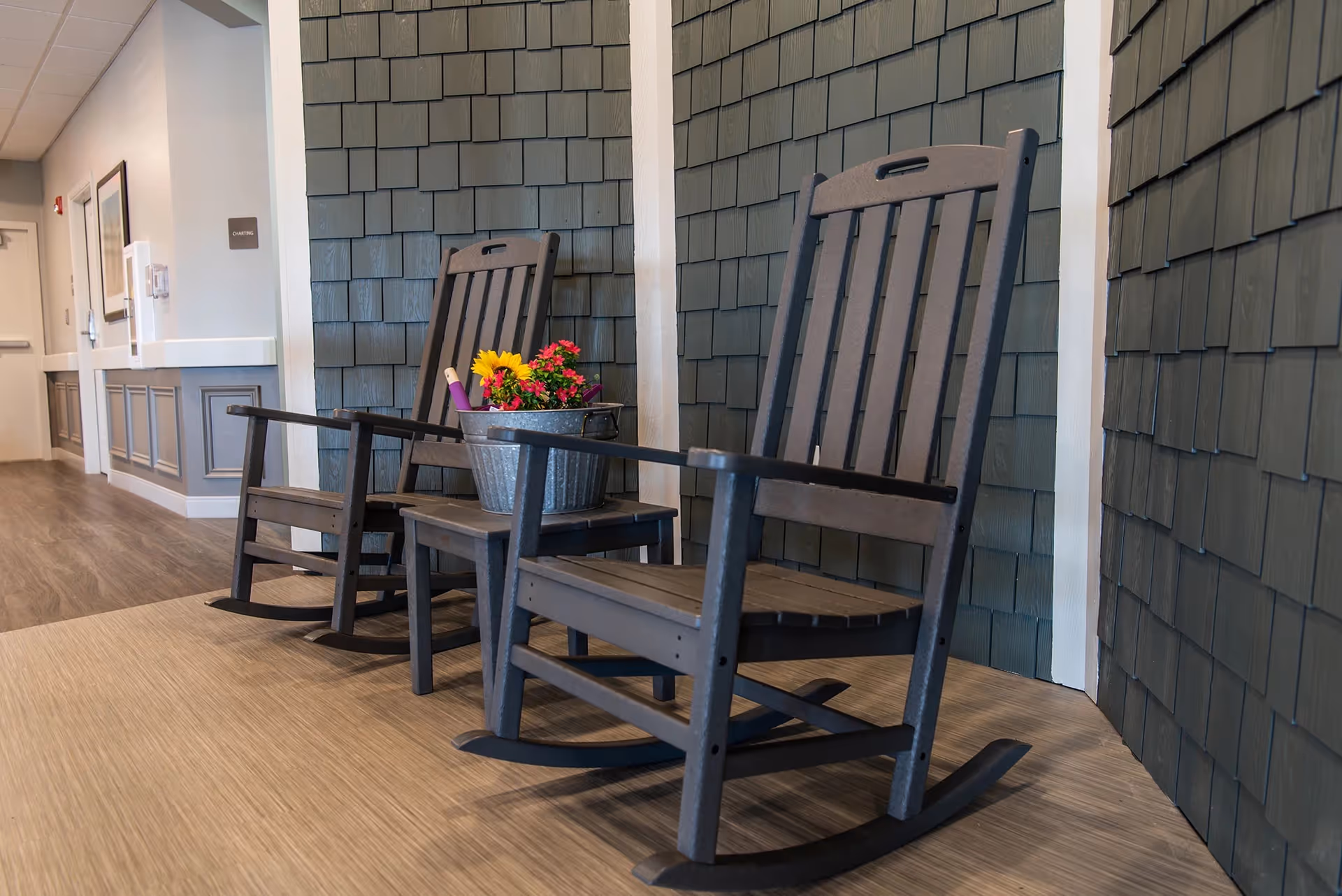Two dark wooden rocking chairs placed on a light wood floor in a hallway with gray shingle walls. A metal bucket with colorful flowers is on one of the chairs. The hallway has light-colored walls and a door at the end.
