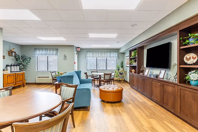 Bright communal living room with sofas, chairs, round tables, a large wall-mounted TV in built-in wooden cabinetry, and wood-look flooring.