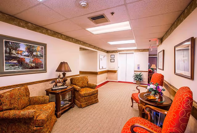 A cozy hallway seating area in a retirement village with two patterned armchairs and a small wooden table with a lamp and decorative sculpture on the left side. On the right side, there are two red upholstered chairs with a round wooden table between them, holding a flower arrangement. The hallway has beige walls with a decorative border near the ceiling, carpeted floor, framed artwork on the walls, and an elevator at the end.
