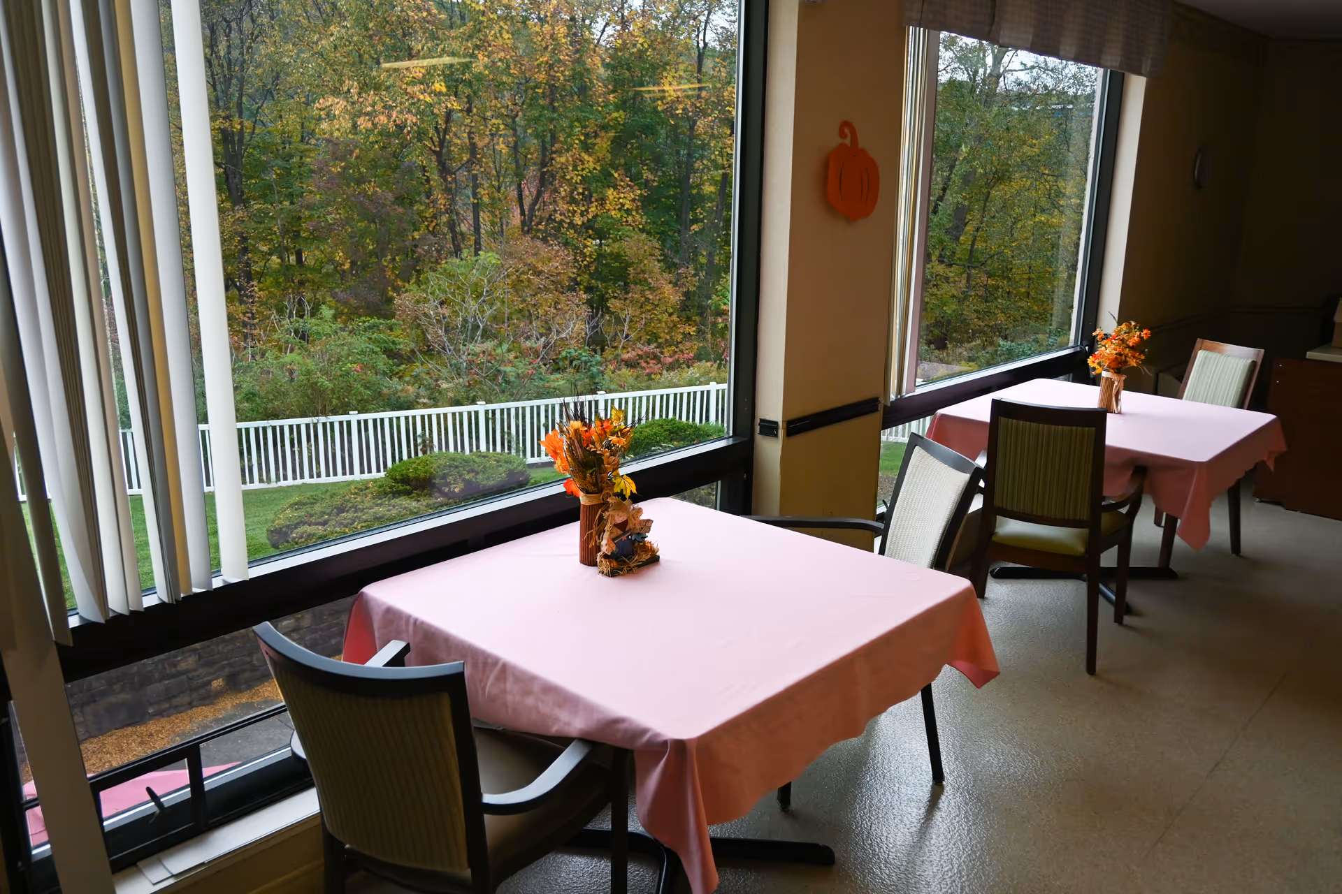 Two dining tables covered with pink tablecloths, each with a small autumn-themed floral centerpiece, positioned next to large windows overlooking a green outdoor area with trees and a white fence.