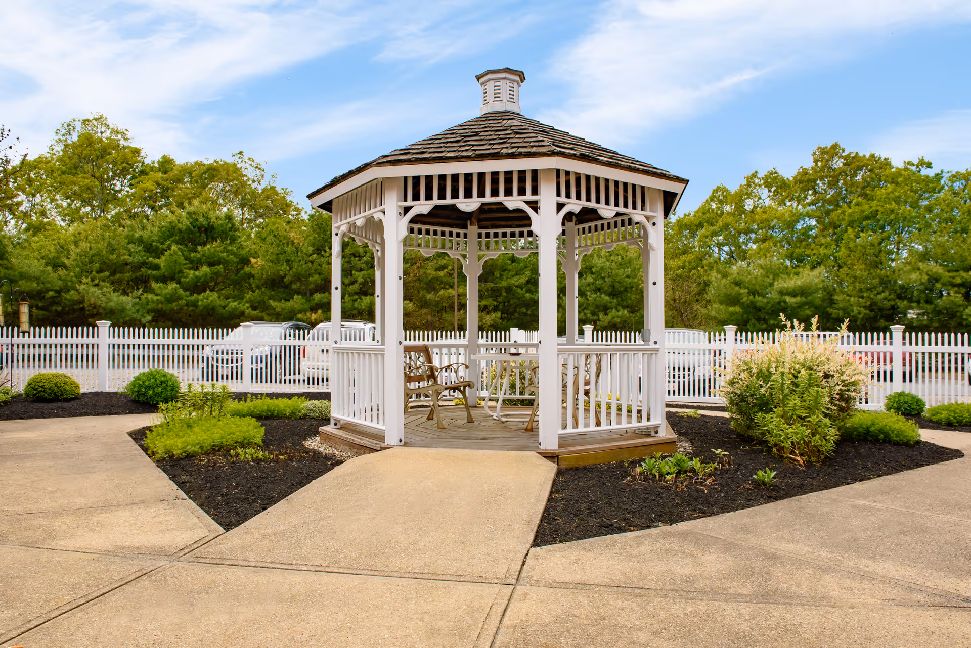 White wooden gazebo on a paved courtyard surrounded by shrubs, a white picket fence, and trees in the background.