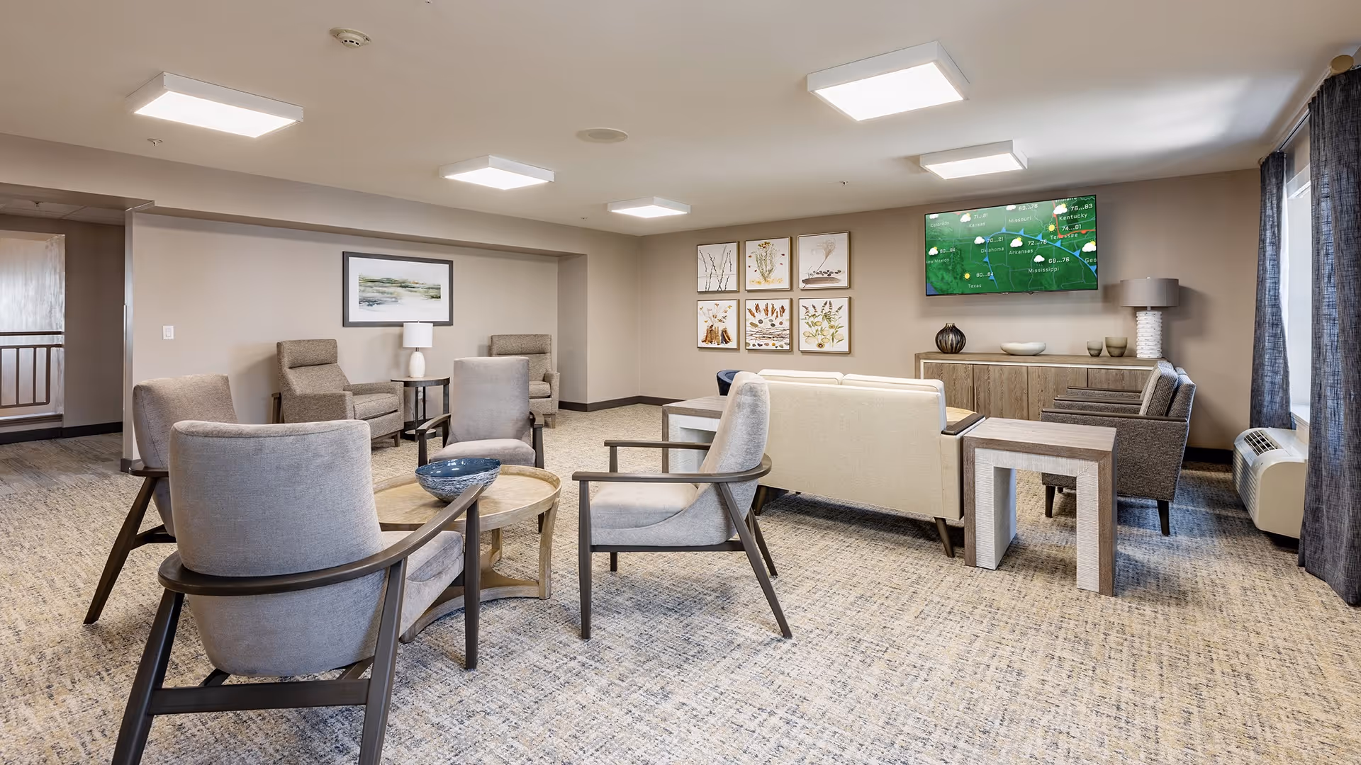 A spacious living room area in a senior living facility with multiple comfortable armchairs and a sofa arranged around a small round wooden table. The room features neutral tones with beige walls, carpeted floor, framed botanical artwork on the wall, a flat-screen TV mounted above a wooden cabinet, and large windows with dark curtains allowing natural light to enter.
