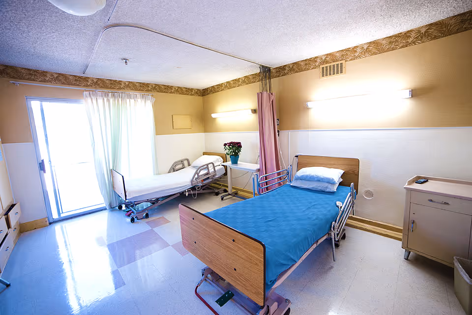 A bright convalescent patient room with two adjustable hospital beds, a bedside cabinet, curtains, and a sliding glass door.