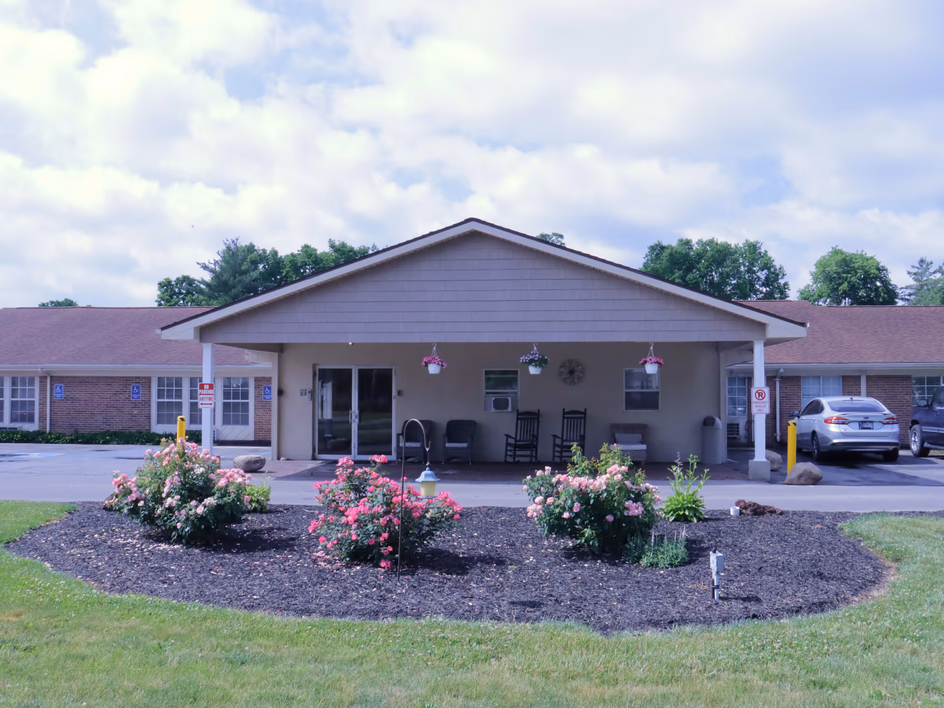 Front entrance of a single-story senior care building with a covered portico, rocking chairs, parked cars, and flower beds in the foreground.