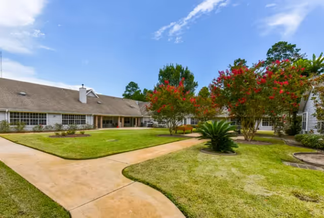 A landscaped outdoor courtyard area with green grass, paved walkways, and flowering trees surrounded by a single-story building under a partly cloudy sky.