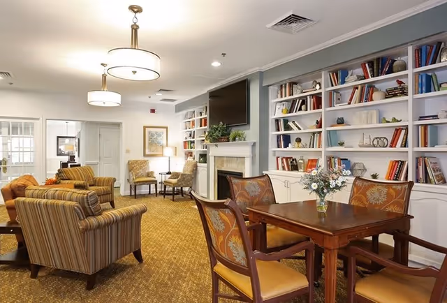 A cozy living room area with striped armchairs, a wooden table with four chairs, a fireplace with a TV mounted above it, and built-in bookshelves filled with books and decorative items. The room is well-lit with ceiling lights and has a carpeted floor.