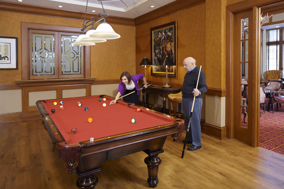 An elderly man and a younger woman playing pool on a red felt pool table in a warmly decorated room with wooden floors and walls. The woman is leaning over the table aiming her shot while the man stands nearby holding a cue stick. The room features framed artwork, a lamp on a side table, and a doorway leading to another room with red carpet and chairs.