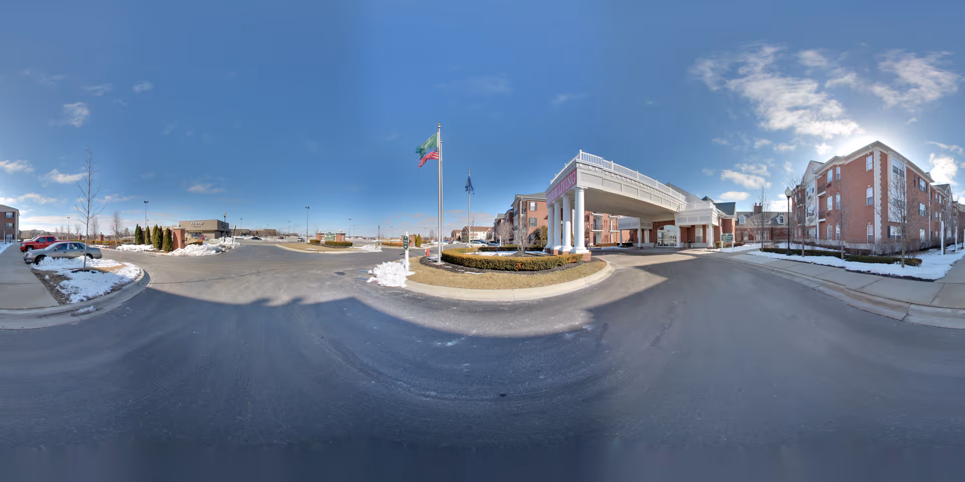 Wide exterior view of Waltonwood Lakeside senior living facility showing a large driveway with a covered entrance supported by white columns, surrounding brick buildings, a flagpole with flags, some parked cars, and patches of snow on the ground under a clear blue sky.