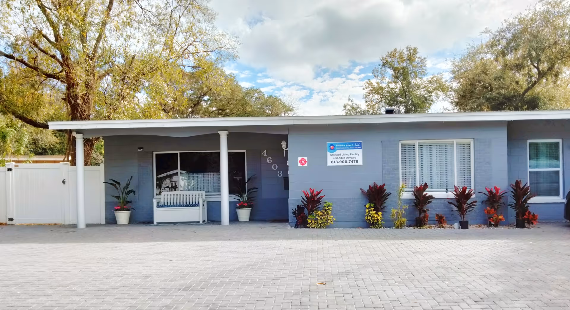 Front exterior view of a single-story assisted living facility building with a covered porch, white bench, potted plants, and a sign displaying the facility's name and contact number. The building is painted gray with white trim and surrounded by trees and a paved driveway.