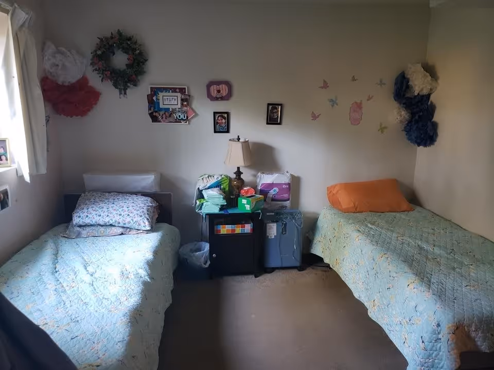 A small bedroom with two single beds on opposite sides of the room. Each bed has a quilted bedspread, one with a floral pillow and the other with an orange pillow. Between the beds is a small black nightstand with a lamp, various supplies, and a trash bin underneath. The walls are decorated with framed photos, a wreath, paper flowers, and butterfly stickers. A window with white curtains is partially visible on the left side.