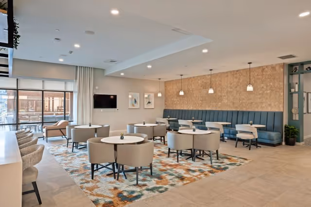 A modern dining area with round tables and cushioned chairs arranged on a colorful patterned rug. There is a long blue banquette seating along a textured wall with hanging pendant lights above. Large windows let in natural light, and a television is mounted on one wall. The space has a clean, bright, and welcoming atmosphere.