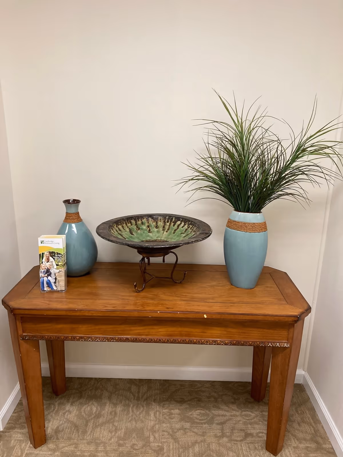 A wooden console table against a beige wall with a decorative green and brown bowl on a metal stand in the center. To the left is a blue vase with a brown band near the top and a brochure holder with Cambridge Retirement Living brochures. To the right is a taller blue vase with a brown band near the top, holding green ornamental grass.