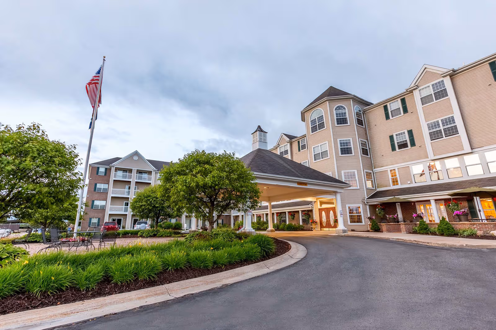 Exterior view of Independence Village Of Petoskey, showing a multi-story beige building with green shutters, a covered entrance with columns, landscaped greenery, trees, and an American flag on a flagpole.