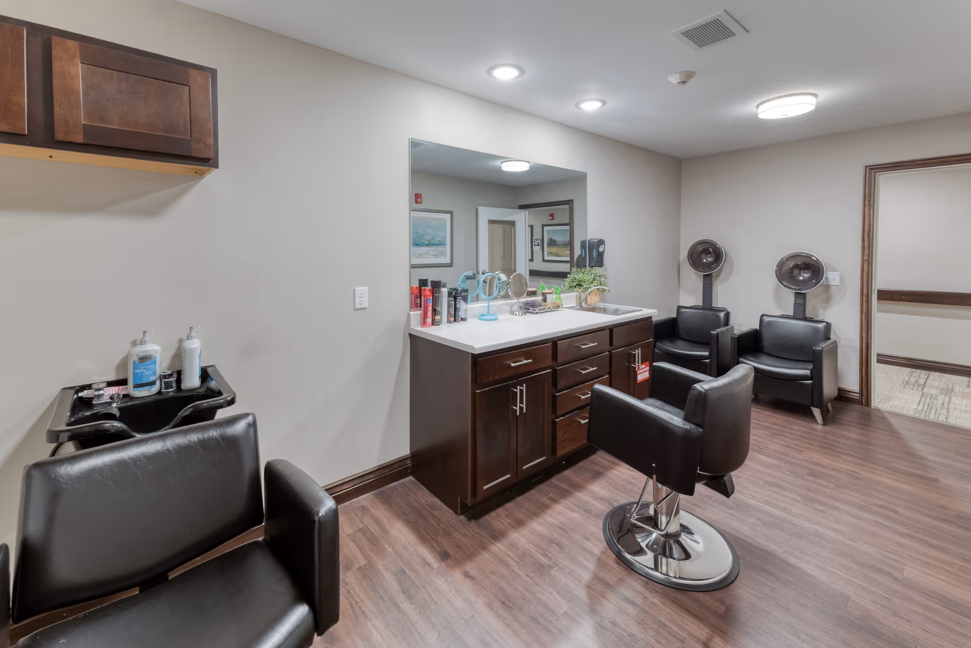 Interior of a senior living facility hair salon with black salon chairs, hair dryers, a large mirror above a countertop with hair products, and a small sink for washing hair. The room has wood flooring and neutral-colored walls.