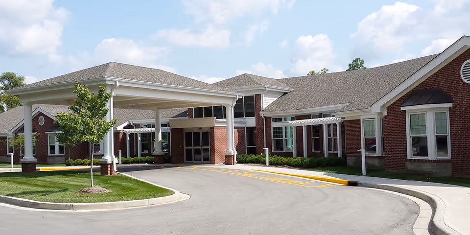 Front entrance of a brick senior living building with a covered drive-up portico, driveway, and landscaped lawn.