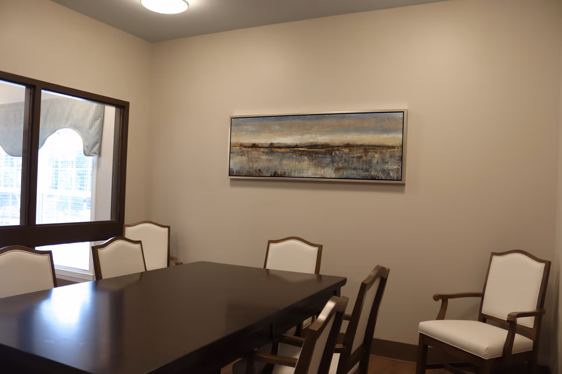 A dining room with a dark wooden table surrounded by six white cushioned chairs with wooden frames. There is a large window with a valance on the left wall and a landscape painting hanging on the beige wall.