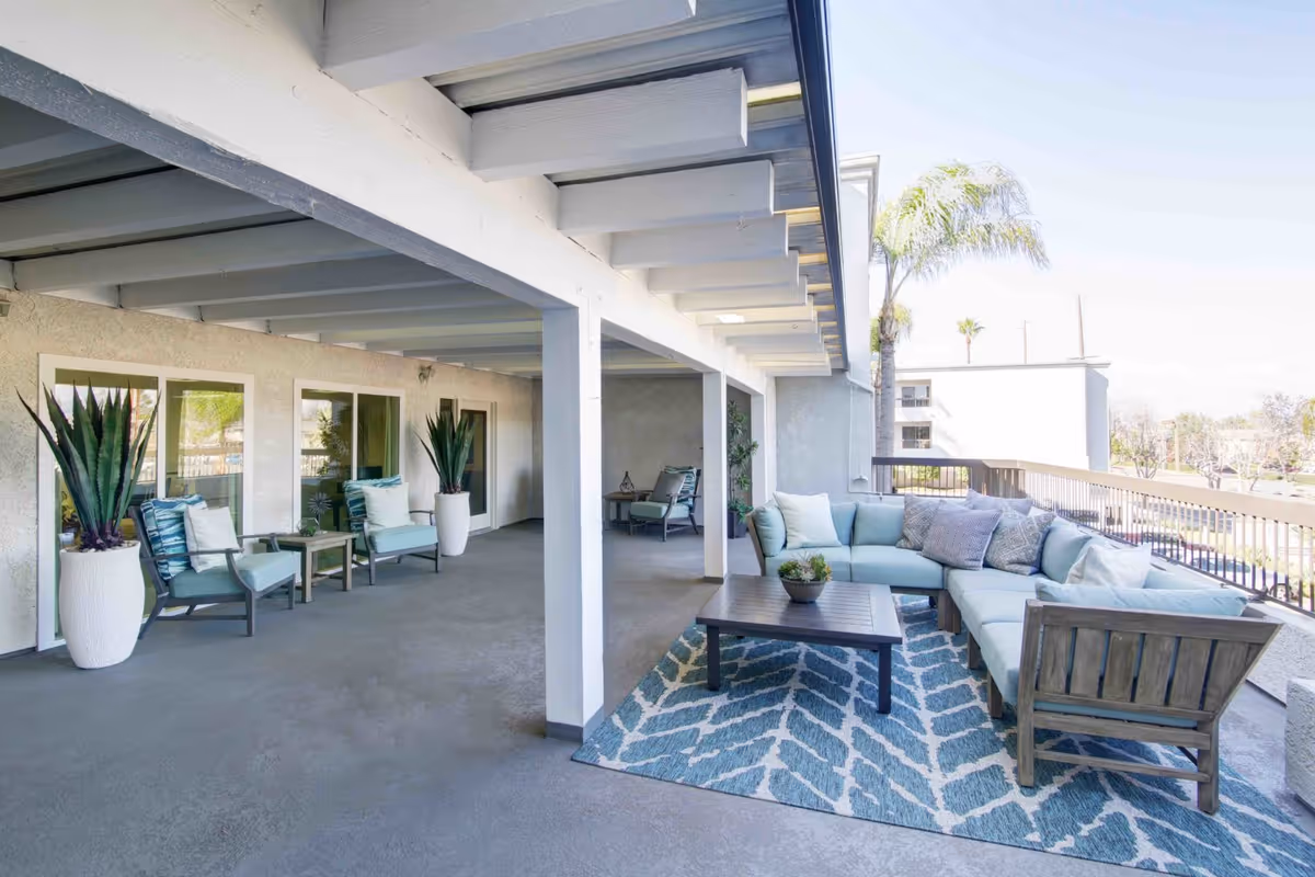 Spacious covered outdoor patio area with light blue cushioned sectional sofa on a patterned blue and white rug, wooden coffee table with a small plant, additional seating with two armchairs and side table, large potted plants, and a view of palm trees and buildings in the background.