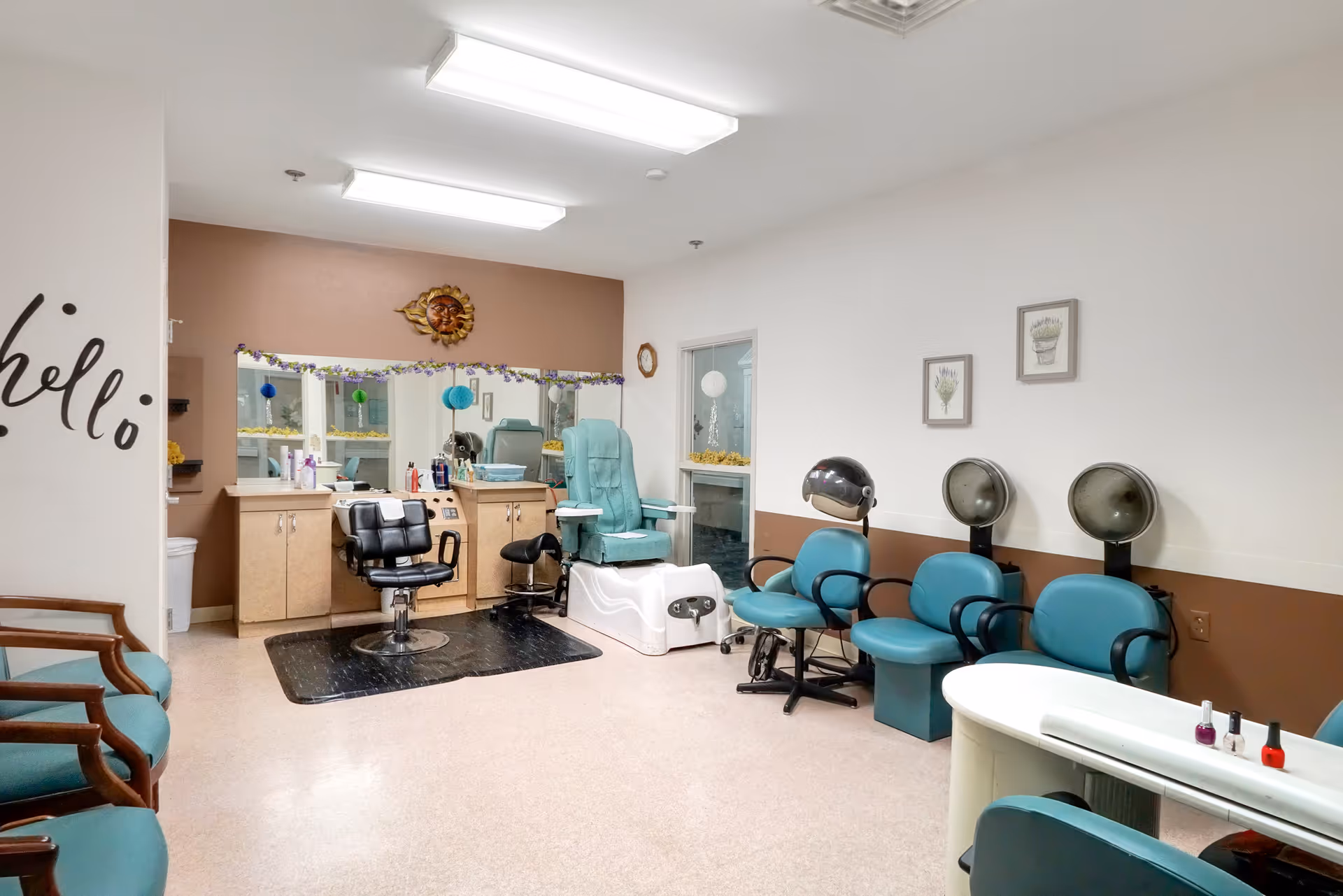 Interior view of a salon area in a senior living facility with salon chairs, hair dryers, a pedicure chair, and a manicure table. The walls are painted white and brown, with decorative wall art and a 'hello' decal visible.