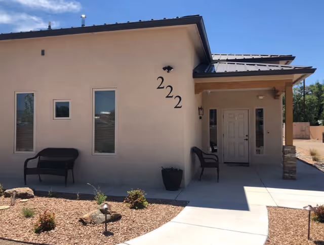 Exterior view of a single-story building with beige stucco walls and a metal roof. The building has three windows and a white front door under a covered porch supported by wooden beams and stone pillars. Two black benches are placed outside near the windows and the entrance. The building number '222' is displayed on the wall near the door. The surrounding area features a landscaped garden with rocks, small plants, and a concrete walkway leading to the entrance. The sky is clear and blue.