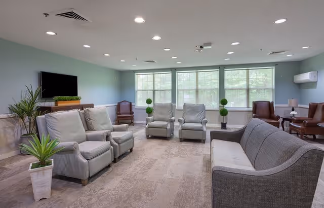 A spacious living room with multiple armchairs and a sofa arranged around the room. Large windows allow natural light to fill the space. There is a flat-screen TV mounted on the wall, several potted plants, and soft lighting from ceiling fixtures.
