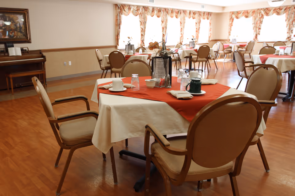 A dining room with several tables covered with white tablecloths and red table runners. Each table is set with cups, glasses, napkins, and a centerpiece. The room has wooden flooring, beige walls, and large windows with patterned curtains allowing natural light to fill the space. A piano and a framed picture are visible in the background.