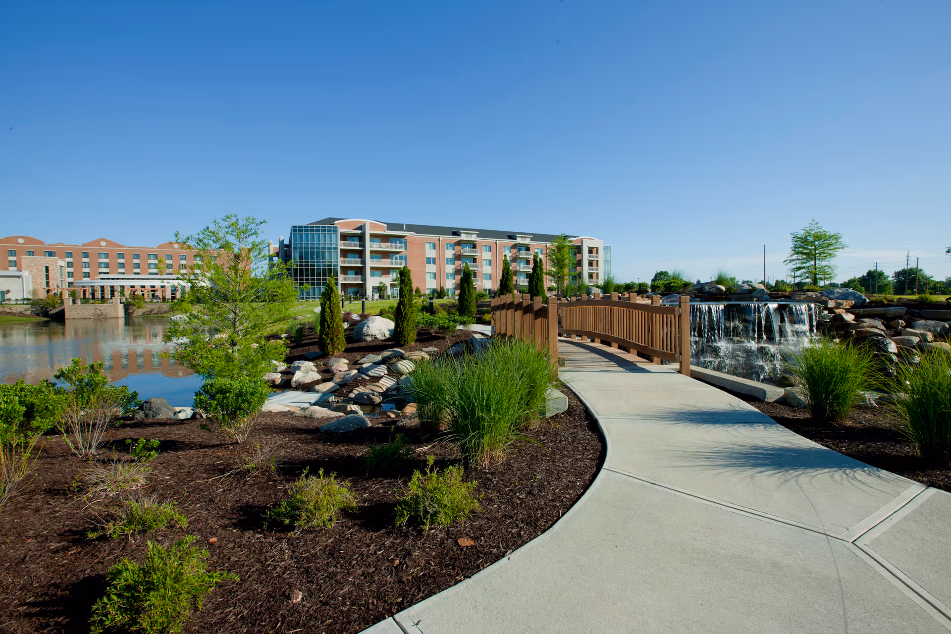 A landscaped outdoor area with a concrete pathway leading to a wooden bridge over a small waterfall feature. There are shrubs, small trees, and rocks along the path, with a large multi-story brick building in the background under a clear blue sky.