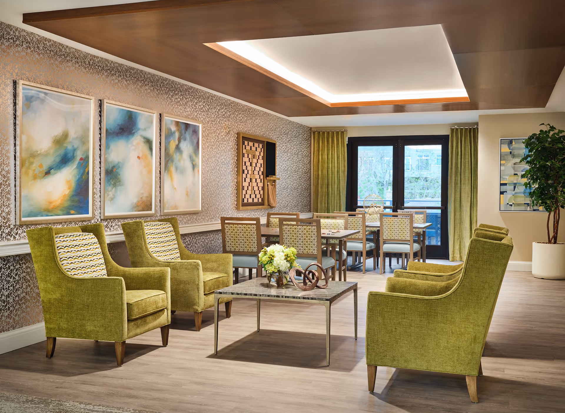 A cozy and well-lit common area in an assisted living facility featuring four green upholstered armchairs arranged around a square coffee table with a flower arrangement and decorative sculpture. In the background, there are two tables with chairs, a large window with green curtains, abstract artwork on the walls, and a chessboard mounted on the wall.