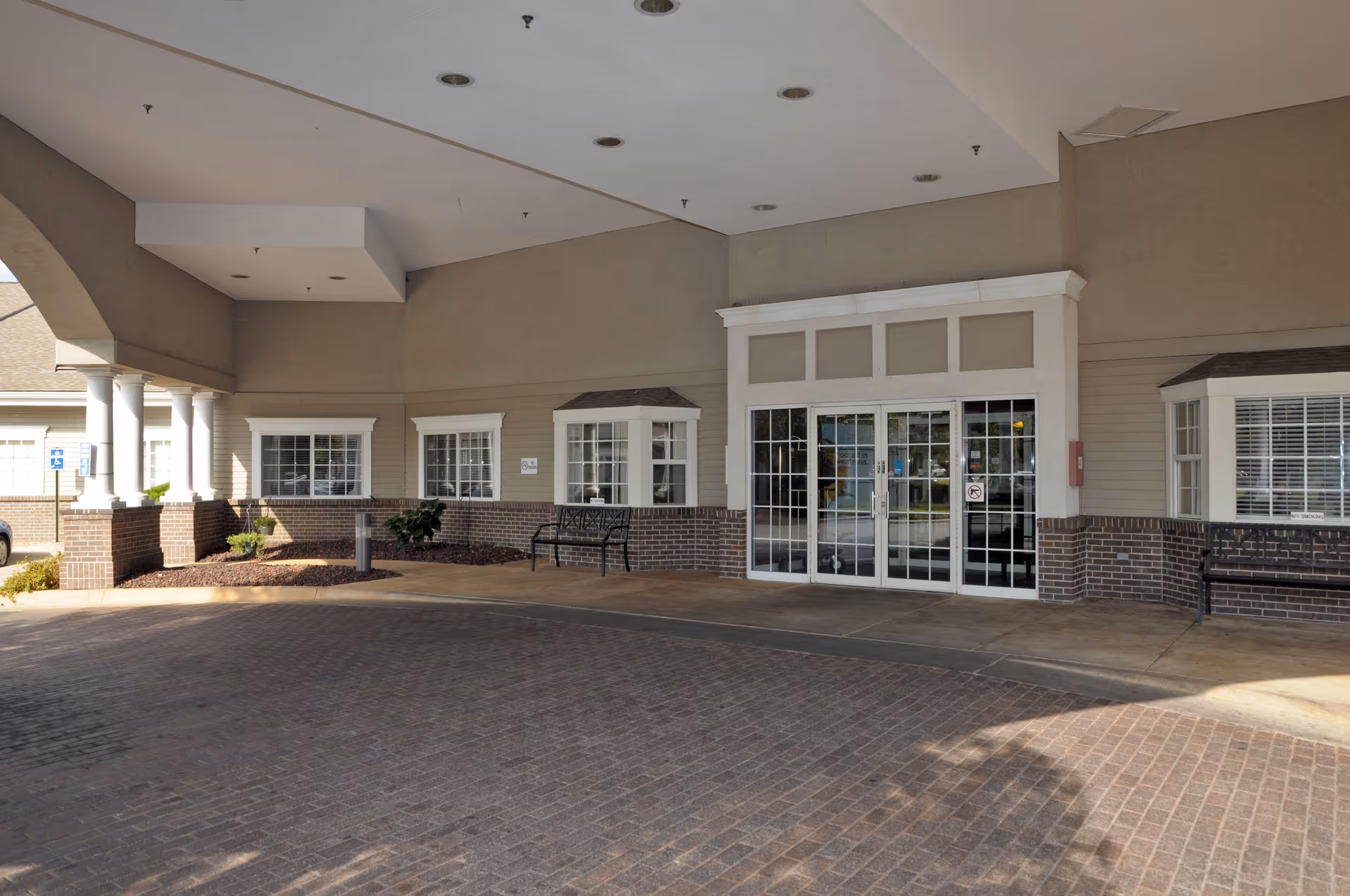 Covered porte-cochere entrance with glass double doors, benches, columns and brickwork at a senior living facility.