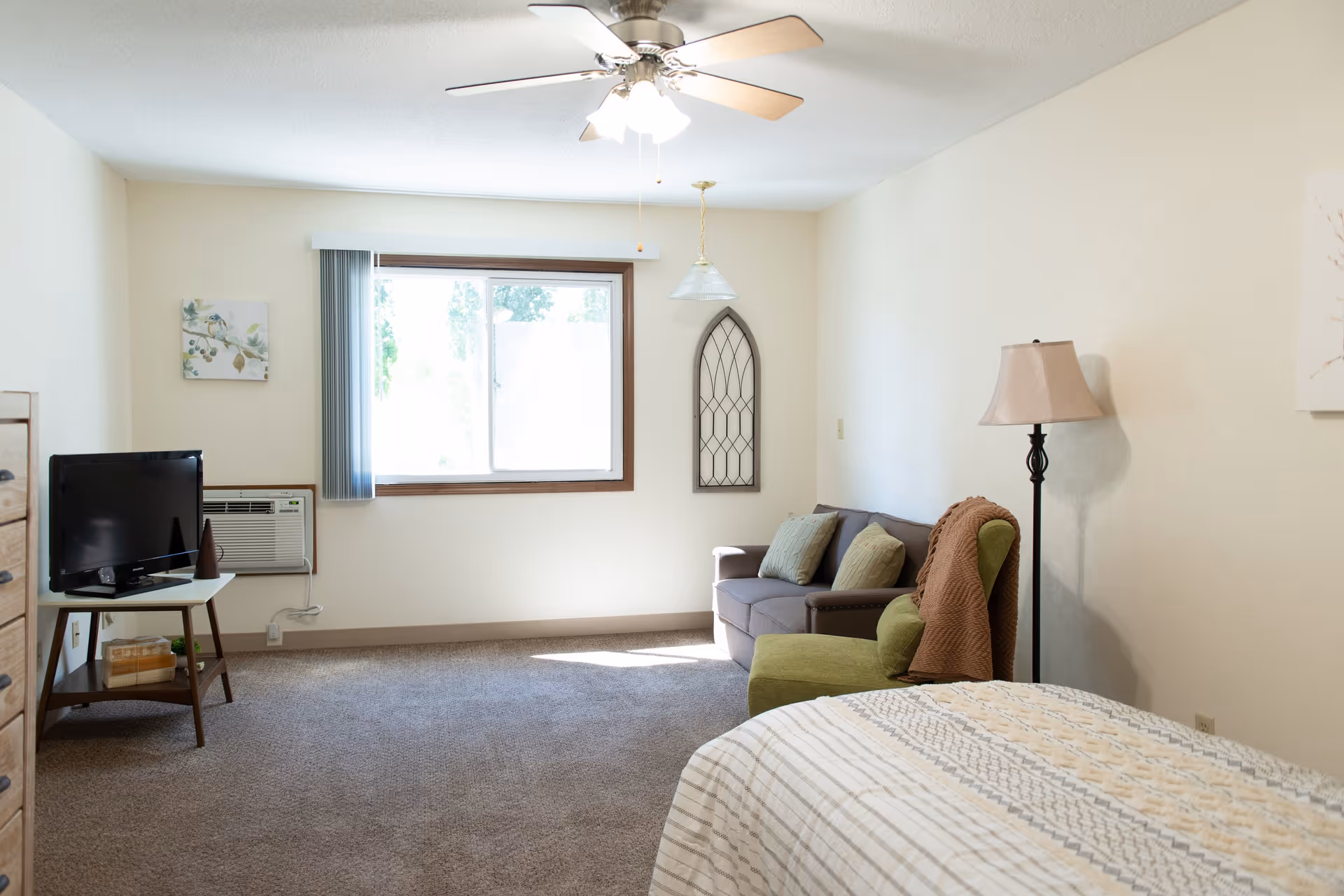 A bright and cozy bedroom with a bed in the foreground, a green armchair with a brown throw blanket, a gray couch with two pillows, a floor lamp, a small TV on a stand, and a window with vertical blinds letting in natural light.
