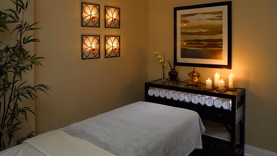 A serene massage room with a massage table covered in a light gray sheet, a black console table holding rolled white towels, lit candles, a small statue, and a framed picture on the wall. Four wall-mounted candle holders with lit candles and a potted plant are also visible.