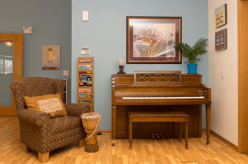 Cozy corner of a living room with a patterned armchair, a small drum, a wooden upright piano with a matching bench, a framed floral painting above the piano, a potted plant on top of the piano, and a magazine rack filled with magazines against a light blue wall.