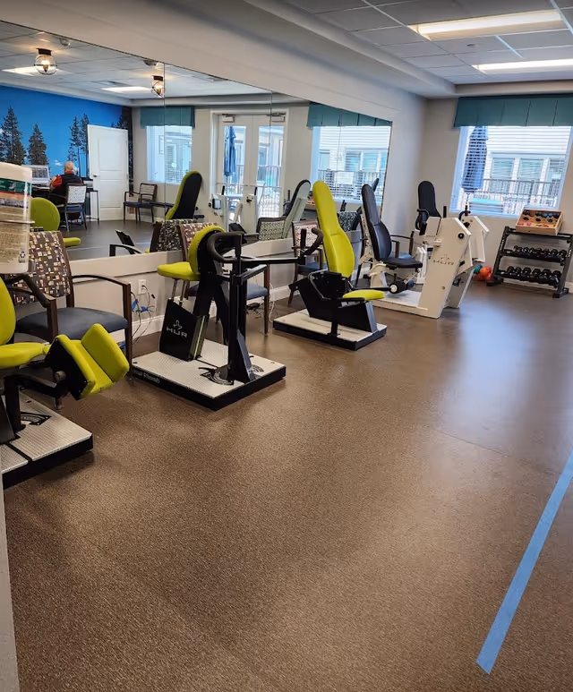 Bright interior fitness/therapy room with several exercise machines with green seats, mirrors on the wall, chairs and a rack of free weights by the windows.
