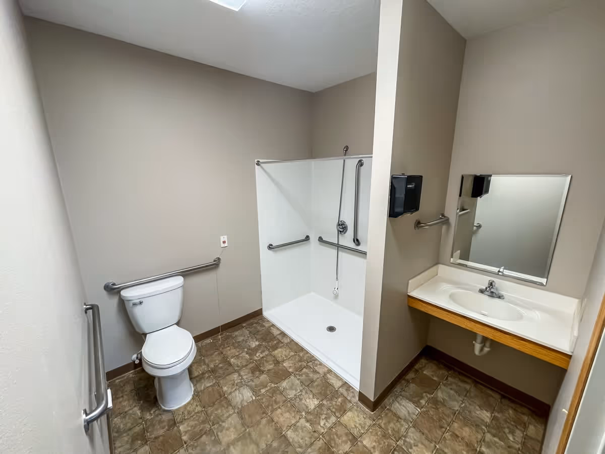 A clean, accessible bathroom with beige walls and tiled floor. The room features a white toilet with grab bars on the walls nearby, a walk-in shower with multiple grab bars and a handheld showerhead, and a white sink with a mirror above it and a wall-mounted soap dispenser.