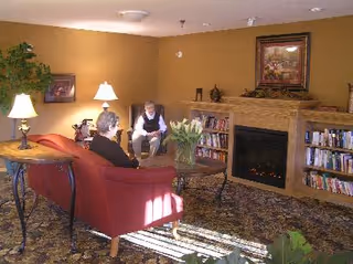 A cozy living room with a red sofa, a coffee table, and a fireplace. Two people are seated, one reading a book, with bookshelves in the background and warm lighting.